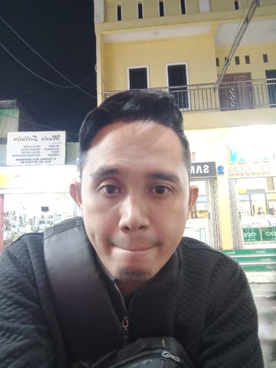 A male individual, likely of Southeast Asian ethnicity, is depicted in a close-up, head-and-torso shot, centered and facing the camera with a neutral expression. He possesses short dark hair styled with a pompadour and a short goatee. He is wearing a dark, quilted zip-up jacket, and a dark bag strap is visible over his left shoulder, with a dark pouch resting in the lower foreground.
The scene is illuminated by artificial light at night in Sarimulyo, Indonesia. In the background, a two-story building with a light-yellow facade is visible. The upper floor features narrow vertical windows and a balcony with a metal railing. The ground floor displays brightly lit commercial storefronts. Reversed text on prominent signs includes "Muda Selluler," "SAMSUNG," and "OPPO," indicating mobile phone or electronics retailers. One individual is dimly visible inside one of the stores. Electrical wires traverse the dark night sky in the upper left portion of the image.