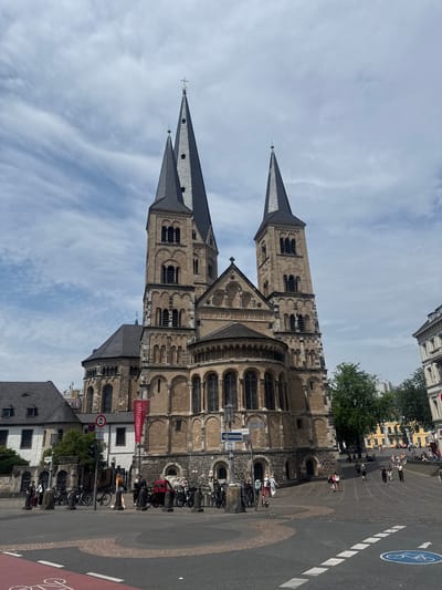 The image shows the Bonn Minster, a Romanesque basilica in Bonn, Germany. The church is large and made of stone, with two tall spires. In front of the church, there is a public square with people walking and standing around. There are also many bicycles parked in racks. The sky is partly cloudy. The photo appears to be taken on a pleasant day, with good visibility and natural lighting.