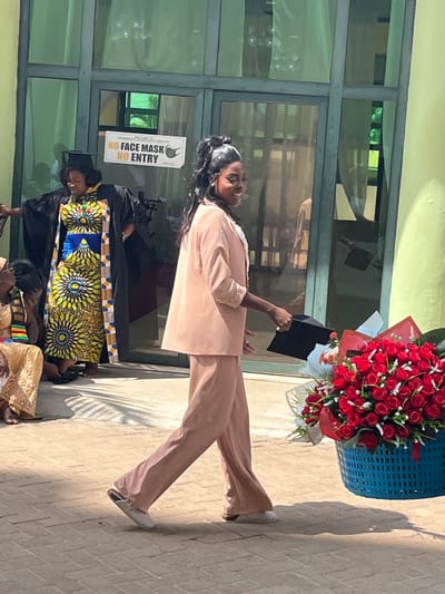 This image depicts an outdoor celebratory scene, likely a graduation, in Madina, Ghana. The primary subject is a young Black woman in the mid-foreground, walking from left to right with a wide smile. She wears a light peach blazer over a white top with pearl-like embellishments, matching wide-leg trousers, and light-colored flat mules. In her left hand, she holds a black academic cap. To her immediate right, on the ground, a large bouquet of red roses, accented with small white flowers and green foliage, rests in a blue plastic basket.
In the background, positioned near a building entrance, a Black woman stands wearing a black academic gown over a vibrant yellow and blue patterned traditional dress, topped with a black mortarboard. Her hands are slightly extended. To her left, partially visible, another person is seated, adorned in yellow traditional fabric and a patterned sash. The building behind them features large glass-paned doors and windows, with a light green wall section to the far right. A yellow sign affixed to the glass door clearly reads "NO FACE MASK NO ENTRY" with a face mask icon, and below it, "WELCOME TO CHARISMATIC HOUSE MINISTRY". The ground is paved with light-colored interlocking pavers, and bright sunlight casts distinct shadows across the scene, indicating clear weather.