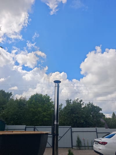 The image depicts an outdoor scene under a blue sky with white clouds. In the foreground, a black structure with a wooden top is visible on the left. A tall, shiny metal chimney stands vertically in the center, emitting a small plume of smoke. Behind the chimney, a grey metal fence runs horizontally across the middle ground, with a dense line of green trees beyond it. To the right, a white car is parked on a brick-paved area. The location is identified as Лужники Снт, Russia.