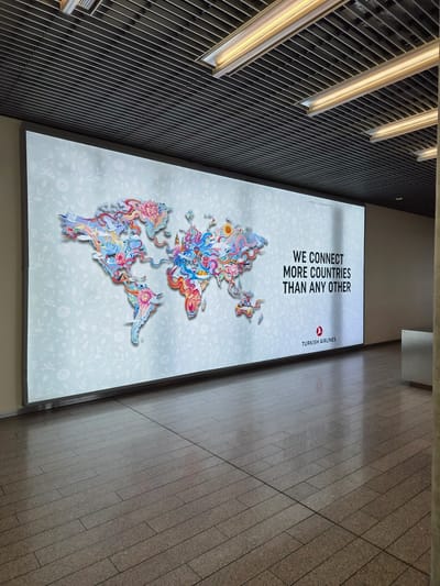 The image depicts an advertisement for Turkish Airlines in what appears to be a modern architectural space. The advertisement is large and illuminated, featuring a stylized world map composed of colorful, swirling patterns and floral designs, set against a light blue backdrop with a repeating flower motif. In bold black text at the top, it reads "WE CONNECT MORE COUNTRIES THAN ANY OTHER" followed by the Turkish Airlines logo.
The advertisement is positioned next to a wall with a dark, linear architectural design and is partially illuminated by strip lighting. The floor is tiled with large rectangular stones.
Given the reference to Niederanven, Luxembourg, it's likely the advertisement is located within Luxembourg Airport (Findel Airport), which is the primary airport serving Luxembourg and located in Niederanven.