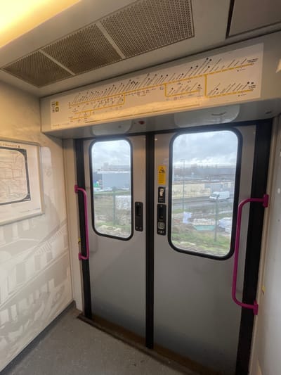 The image depicts the interior of a public transport vehicle, likely a tram or light rail, situated in Gennevilliers, France. This location is confirmed by the route map displayed above the doors, which clearly shows stations such as "Gennevilliers - Les Courtilles" on the T1 tram line, and a broader regional map of Île-de-France on the left wall.
The foreground presents a gray, speckled floor. Central to the frame are two sets of double-leaf doors, light grey with prominent black vertical frames, each featuring a tall, rounded-rectangular window. Recessed vertical handles are visible on the door panels, alongside yellow warning stickers that include a hazard symbol (triangle with exclamation mark) and text likely advising caution. Bright pink/magenta vertical grab handles are mounted on the walls flanking the doors.
Above the doors, a backlit horizontal route map outlines a public transport line with numerous stations. The ceiling is light-colored, featuring a linear light fixture and several rectangular panels with hexagonal perforations, indicative of ventilation or air conditioning grilles, some displaying minor dirt accumulation. The left wall holds a white-framed map of the Île-de-France transport network, and below it, the wall is decorated with a faint, repeating monochrome graphic depicting architectural outlines.
Through the windows, an urban-industrial landscape is visible under an overcast sky. Directly outside, a strip of sparse vegetation and dirt borders a chain-link fence, beyond which lies a road. Piles of construction materials, including bags of aggregate and green tarpaulins, are present in the area adjacent to the tracks. A white commercial van is visible on the road. In the middle and far distance, various industrial and commercial buildings, including a large, grey, flat-roofed structure and taller, multi-story buildings, populate the urban periphery. No individuals are present or visible within the vehicle or in the external environment. No specific actions or interactions are depicted.