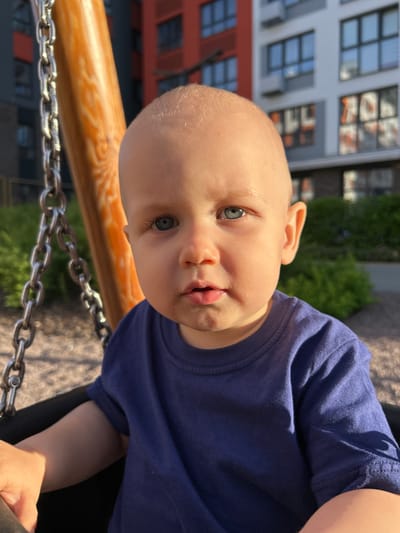 A close-up, vertically oriented shot features a baby with light skin and blue eyes. The baby is wearing a dark blue t-shirt and is looking upwards towards the camera. The baby's head is tilted, and their mouth is slightly open. In the foreground, a wooden bar is visible, with a metal chain running beneath it. The background is blurred, but suggests an outdoor setting with a building that has many windows and green foliage. The lighting suggests it is daytime, and the weather appears to be clear. The image appears to be taken at a playground, possibly in Kaluga, Russia. There is no visible text in the image.