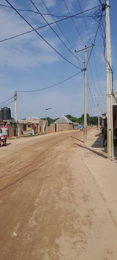 This image depicts a street scene in Zanzi, Nigeria, characterized by a dirt road and buildings constructed from concrete blocks and corrugated metal. The sky is blue with scattered clouds. Power lines are prominently featured, stretching across the frame, supported by concrete poles.

On the left side of the road, a man is pushing a wheelbarrow containing various items. Adjacent to him, a building with a water tank on top and barbed wire fencing is visible. Further down the street, several more structures with distinct architectural styles, including one with a conical metal roof, line the road.

In the mid-ground, a yellow motorized rickshaw is moving down the street, and other individuals are walking in the distance. The right side of the road features a covered walkway or shopfront, with a few people standing or sitting nearby. The overall impression is of a developing urban environment with basic infrastructure.
