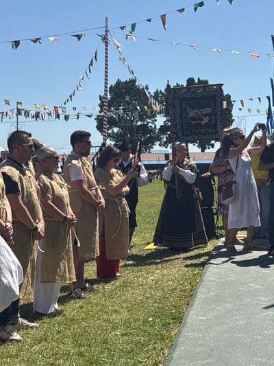 The image depicts an outdoor gathering with a clear blue sky and a line of people. Several individuals are wearing rustic, brown tunics tied with rope, possibly representing a historical or cultural theme. In the background, a banner with "ASOCIACION CULTURAL LA MADRENA" is visible, suggesting a local organization's event. Decorative flags are strung across the scene, and a tall, striped pole stands prominently. One woman in the foreground is holding a banner and appears to be leading a procession or demonstration. Other attendees are observing, some taking photographs with their mobile phones. The ground is covered in green grass, and a paved pathway is also present. The scene suggests a festive or ceremonial occasion taking place in Llanera, Spain, as indicated by the presence of the "La Madrena" cultural association.