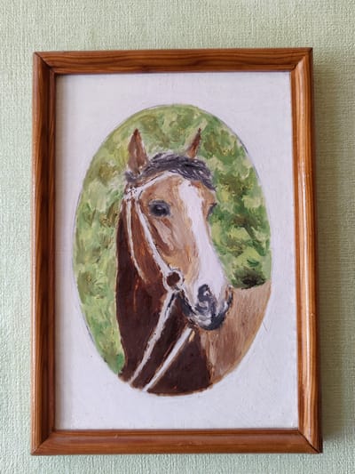 A framed painting of a horse's head is displayed on a pale green textured wall. The painting is rendered in an oval shape within a rectangular white mat and a wooden frame. The horse, depicted in shades of brown and white, faces towards the viewer with a direct gaze. It wears a simple bridle. The background within the oval is a brushy green, suggesting foliage. The overall style of the painting appears to be expressive and somewhat impressionistic. The lighting suggests an indoor setting, with no indication of time of day or weather. There is no visible text in the image. The location context of Rybnitsa, Moldova, does not provide any visual cues within the image itself.