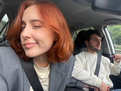 Two people are inside a car. A fair-skinned woman with red hair and wearing a seatbelt is in the passenger seat. She wears a gray blazer over a light-colored t-shirt, and a silver chain is visible around her neck. She is partially facing the camera. Next to her, a fair-skinned man with dark hair and a beard is in the driver's seat, also wearing a seatbelt. He is wearing a white sweater. He is holding a blue and white object in his hand. The car appears to be driving on a road with green foliage visible through the windows. This image does not depict the city of Grigalati, Georgia.