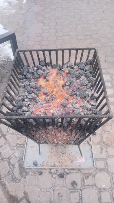 A square metal fire pit grill is filled with glowing, burning charcoal. The intense heat radiates from the center, where flames are visible amidst the dark grey rocks. The grill sits on a grey metal tray, positioned on a paved surface of interlocking stones. A small amount of ash and unburned charcoal surrounds the grill.