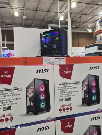 The image shows a retail environment, likely a warehouse or large store, with industrial lighting and exposed ceiling infrastructure. In the foreground, there are stacked boxes for "MSI AEGIS GAMING DESKTOP" PCs. The boxes display specifications such as "PRO PERFORMANCE GAMING PC," "AMD RYZEN™ 9 9900X," and "NVIDIA® GEFORCE RTX™ 5080." The top box has a price tag of "$3199.99" and item number "1927586". To the left of the MSI boxes, a partially visible "Galaxy S20+ 5G" label is present. Further back, a black gaming computer tower with RGB lighting is visible on a shelf. The overall scene suggests a commercial setting where electronics, specifically high-performance gaming computers, are being sold. This scene appears to be located at a Costco store in Wilsonville, Oregon, United States of America, based on the common presence of such displays and the specific product labeling.