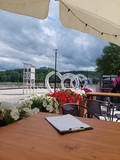 This image shows a view from an outdoor cafe table in Alikonovka, Russia, with a sandy beach and a lake in the background. A wooden table in the foreground holds a clipboard with blank white paper. Behind the table, there are several outdoor chairs, some of which are partially obscured by planters filled with white and red petunias. In the mid-ground, a white lifeguard tower stands on the beach. Further back, large white circular decorative structures are visible, along with a lake with a few boats and people. The background features a treed hill with buildings on top, and a cloudy sky. A person in a pink top and backpack is visible in the far right background.