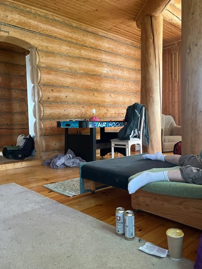 The image shows an interior scene with wooden walls and floors, potentially a cabin or house. A person is sitting on a green couch, wearing shorts and white socks. Next to the couch, there are two beer cans, a disposable coffee cup, and a piece of paper. An air hockey table is positioned against the wooden wall, along with a white chair and a dark duffel bag. There are no specific indicators within the image to confirm that the location is the city of Пятница, Russia.