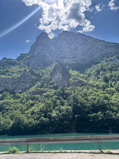 This image depicts a mountainous landscape in Glogošnica, Bosnia and Herzegovina, under bright daylight conditions. The scene is dominated by a large, verdant mountain range on the left and a layered rock face on the right, both rising prominently from a body of turquoise-green water in the foreground.
The upper portion of the image features a clear blue sky with scattered white cumulus clouds. A strong sunbeam or lens flare emanates from the upper-left, extending diagonally across the sky.
The mountains are characterized by steep, rugged slopes. The left mountain is extensively covered in dense green deciduous trees, while the right mountain face displays prominent horizontal geological stratification, revealing distinct layers of rock. Patches of trees and vegetation are also visible on the rocky ledges and crevices of the mountains.
In the lower-middle section, a calm body of water, likely a river, stretches horizontally, exhibiting a distinct turquoise-emerald hue.
A metallic guardrail runs across the immediate foreground, positioned between the viewer's perspective and the water. The guardrail is grey and shows signs of graffiti, including the legible inscription "75.09.2000" and other indiscernible markings. The ground beneath the guardrail is gravelly, with sparse patches of green grass and a few small yellow flowers.
The overall lighting suggests a sunny day, likely midday or early afternoon, with strong illumination casting highlights on the clouds and water, and creating a soft haziness over the distant mountain peaks. No people or animals are visible in the scene, and no explicit actions are depicted.