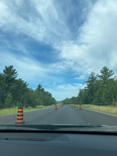 The image captures a view from a vehicle traveling on a rural highway through a forested area in the city of The Nation, Canada. The road ahead is lined with orange and black traffic cones, indicating ongoing construction or maintenance. Dense green trees form a natural barrier on both sides of the asphalt road, with a strip of dry grass and wildflowers running along the shoulder. The sky above is a vibrant blue, crisscrossed with wispy white clouds, suggesting fair weather. The foreground shows the dashboard of the vehicle, slightly out of focus. The scene is peaceful and evokes a sense of a journey through a natural landscape with temporary roadwork.