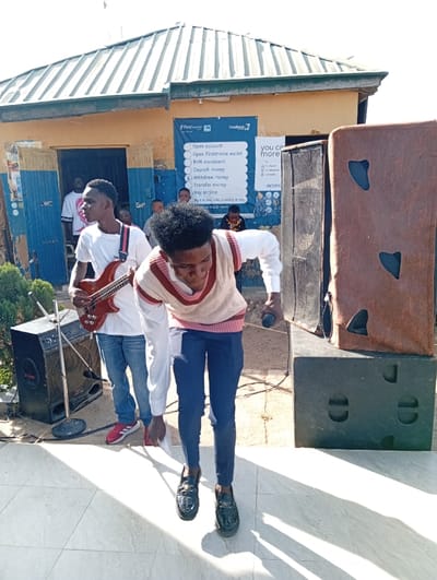 An outdoor scene in Jebbu Miango, Nigeria, depicts two young adult males performing or preparing to perform in front of a single-story building. The male in the foreground, center-right, is leaning forward, holding a black microphone in his right hand. He wears a cream-colored sweater with horizontal red and pink stripes over a white long-sleeved shirt, dark blue slim-fit trousers, and black patterned loafers. To his left and slightly behind, a second male stands, holding a red bass guitar with a white pickguard. He wears a white short-sleeved t-shirt, blue jeans, and red athletic shoes.
Musical equipment includes a large black speaker cabinet on a stand to the left, a microphone stand positioned behind it, and another large black speaker cabinet on the ground to the right, topped with a brown, possibly padded, object featuring irregular cutouts. Cables are visible on the ground connecting the equipment.
The building behind them features light brown/ochre walls with blue trim around an open doorway on the left. Through this doorway, several individuals are partially visible, observing the scene. A corrugated metal roof covers the structure. A blue banner is affixed to the building's facade, displaying "First Bank" and "Firstmonie" logos and listing services such as "Open account," "Open Firstmonie wallet," "BVN enrolment," "Deposit money," "Withdraw money," "Transfer money," and "Buy airtime."
The immediate foreground features a light-colored tiled surface, casting strong shadows from the bright sunlight originating from the upper right. Beyond the tiled area, the ground appears to be dirt or gravel. A small green shrub is visible on the far left.