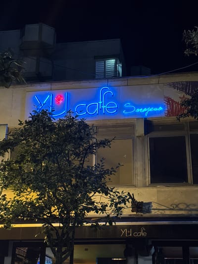 This image shows a neon sign for "YU cafe Sarajevo" on the facade of a building at night. The sign is blue and features a red star. A tree with dense green foliage partially obscures the sign. The building appears to be a commercial establishment, with windows visible on the upper floor. Below the neon sign, a darker awning displays the same "YU cafe" logo in white lettering. The overall environment is dark, suggesting nighttime, with artificial light illuminating the building and signage. The location is identified as Ilidža, Bosnia and Herzegovina.