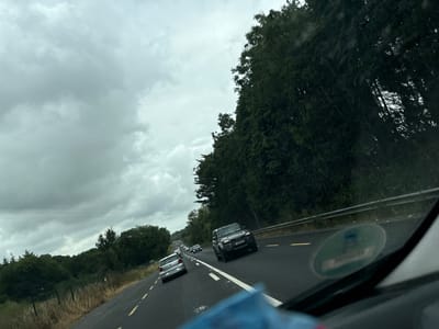 A grey, overcast sky looms over a rural road in Scartnamore, Ireland. The road, marked with yellow lines, curves slightly to the right. Several vehicles are traveling on the road, including a silver Volkswagen Polo in the foreground on the left, a dark SUV with a license plate reading "252-D-379" in the right lane, and other cars further ahead. Dense trees line the right side of the road, with a guardrail separating the road from the trees. The left side of the road features a grassy verge and more trees in the distance. The overall impression is of a slightly dreary day on a country road.
