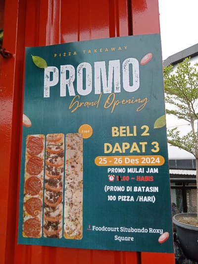 A vibrant promotional poster for a pizza takeaway is displayed vertically on a bright orange corrugated metal surface, likely an exterior wall or food stall. The poster, designed with a teal-green background, announces a "Grand Opening" "PROMO."
The main offer, prominently displayed in white and orange text, states "BELI 2 DAPAT 3" (Buy 2 Get 3 Free), with a "Free" bubble pointing to the offer. The promotion is valid from "25 - 26 Des 2024" (December 25-26, 2024). It starts at "11.00 - HABIS" (11:00 AM - until sold out) and is limited to "100 PIZZA / HARI" (100 pizzas/day).
On the left side of the poster, three rectangular pizzas are depicted: one with pepperoni, another with mixed toppings like minced meat and sliced tomatoes, and a third with a white base and herbs, possibly a focaccia-style or cheese pizza. The poster is adorned with graphic elements such as green leaves and red tomato slices.
At the bottom right, the location is specified as "Foodcourt Situbondo Roxy Square." The background partially visible beyond the poster on the right includes part of a modern building and a large potted plant with green leaves, suggesting an outdoor or open-air food court setting. The bright ambient light indicates it is daytime in Kecamatan Panji, Indonesia.