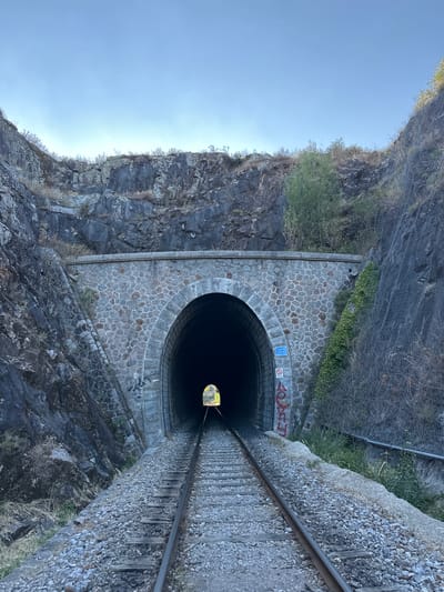 The image shows a view of a stone-arched railway tunnel entrance, a common sight on railway lines in the region, potentially near Générargues, France. The tunnel is set within a rocky, overgrown hillside. Railway tracks run from the foreground towards the dark opening of the tunnel, disappearing into its depths. The tunnel's exterior is constructed of rough-hewn stones, with some ivy growing on the right side. There is graffiti on the tunnel's facade, including red spray-painted lettering and a blue sign with white text. The sky above is clear and blue. The overall scene suggests a remote or rural location, characteristic of the French countryside.