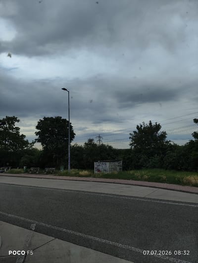 This is a horizontal, eye-level, full-frame photograph taken outdoors on a cloudy day. The scene depicts a paved road in the foreground, with a sidewalk and a grassy area beyond. A solitary streetlamp stands in the middle ground, and behind it, a line of trees and dense foliage forms a backdrop. In the distance, power lines and transmission towers are visible against the overcast sky. The image has a timestamp in the bottom right corner reading "07.07.2026 08:32" and "POCO F5" in the bottom left. The location is identified as Mellingen, Germany. There are no people or animals present in the image.