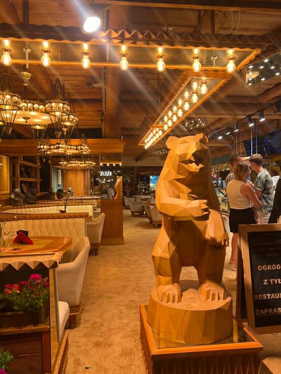 The image depicts the interior of a restaurant in Zakopane, Poland, characterized by a rustic, wooden decor. A large, polygonal wooden sculpture of a bear stands prominently in the foreground, on a wooden pedestal with a mirrored surface. The restaurant features upholstered seating in neutral tones, wooden tables, and decorative lighting fixtures, including chandeliers and exposed bulbs along the wooden ceiling beams. In the background, several people are visible, some interacting at a counter, while others are seated at tables. A blackboard sign with Polish text, likely advertising the restaurant's offerings, is placed next to the bear sculpture. The overall ambiance is warm and inviting, with an emphasis on natural materials and a mountain-inspired theme.