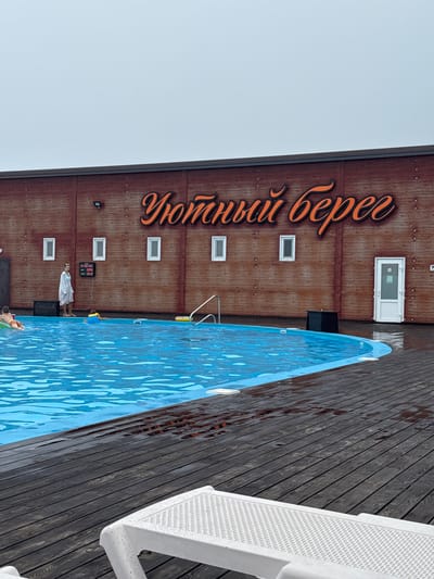 An outdoor swimming pool area at a facility with wooden siding is depicted. The facility has large orange lettering that reads "Уютный берег" (Cozy Shore). A blue swimming pool is in the foreground, with a dark wooden deck surrounding it. Several people are in or near the pool. A woman in a white robe stands on the deck near a digital display. Two children are in the pool, one wearing a yellow floatation device and another on an inflatable ring. In the foreground, a white plastic lounge chair is visible, with water droplets on its surface. The sky is overcast and grey. The location is identified as Rakushka, Russia.
