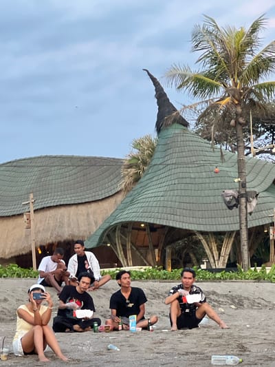 This image depicts a scene at a sandy area in Kerobokan Kelod, Indonesia, likely a beach, during twilight or late afternoon under an overcast sky. Seven individuals are present, six males and one female, all seated on the sand.
In the foreground, a female wearing a white cap, light yellow top, and white shorts is seated with legs crossed, holding a smartphone horizontally in front of her face, seemingly recording or photographing. A disposable plastic cup with a straw is on the sand to her left. To her right, a male in a black t-shirt and black pants is seated, holding an open white food container. A green beverage can is visible near him. Further to the right, a male in a patterned black and white short-sleeve shirt and black shorts is seated, also holding a white food container and a small red packet. An empty plastic water bottle lies on the sand in front of him.
Behind these individuals, on a slightly elevated sandy bank, three more males are seated. The leftmost wears a white t-shirt and dark shorts, looking down at a smartphone. The middle male wears a black t-shirt and is looking forward, with a dark bottle and packaging nearby. The rightmost male wears a white jacket over a black t-shirt and looks towards the right. A seventh male is partially visible further back on the sand.
In the background, two distinctive architectural structures are visible. The prominent building on the right features a green, multi-tiered, conical roof topped with a dark, pointed spire, supported by visible bamboo-like structures at its base. To its left, another building exhibits a wavy, green-shingled roof over a thatched wall structure. A tall palm tree with visible coconuts stands between these two buildings, and two grey bags are suspended from its trunk. A small, reddish-orange object hangs from a wire near the conical roof. Other green foliage is visible behind the sandy bank. The sky is a light blue-grey, and a small, dark, indistinct object is visible high in the sky.