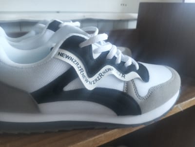 A close-up, angled shot shows a pair of white and black athletic shoes. The shoes are placed on a wooden surface, likely a shelf, with a dark brown, textured object in the background to the right. The shoes are predominantly white with black accents and grey suede detailing on the toe and heel. A white ribbon with black lettering that reads "NEWNZRZRNEWNZRZR" is wrapped around the shoe. The laces are white and tied. The lighting is dim, casting shadows. The scene appears to be indoors.