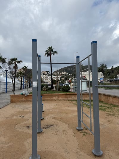 The scene depicts an outdoor calisthenics exercise area in Port D'andratx, Spain, under an overcast sky. In the foreground, a light brown, sandy ground surface features several grey metal calisthenics structures, including vertical posts with horizontal bars and a ladder component, some topped with blue caps.
Behind the exercise area, a low, tan-colored brick retaining wall separates it from a paved pedestrian pathway on the left and a patch of green grass with bushes and small trees on the right. Further back, an asphalt road runs parallel to the wall, where a white van-like vehicle is partially visible. Adjacent to the road, a bus stop shelter with a blank advertising panel stands. A red circular sign, possibly a 'no entry' or 'stop' sign, is visible above the bus stop.
In the midground, several palm trees and deciduous trees line the pathway and are scattered across the landscape. To the far left, white tent-like structures with blue accents are visible behind the pathway. White residential buildings with reddish roofs are situated on a gentle slope in the background, appearing amidst lush green vegetation. A prominent, tall palm tree stands towards the center-left. The background is dominated by a green, tree-covered hillside dotted with more white buildings. The sky is uniformly overcast, exhibiting various shades of grey. No individuals are present in the scene.