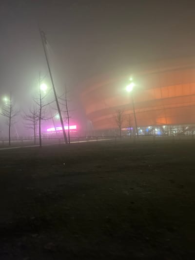 This nighttime photograph depicts an outdoor scene shrouded in heavy fog, likely in Eckbolsheim, France. The prominent subject in the background is a large, modern building featuring a distinctive orange, curved, and ribbed facade, which strongly resembles the Zénith Strasbourg Europe concert hall. Below this main structure, a vivid strip of bright pink/magenta lights cuts through the mist, possibly an illuminated sign or decorative lighting for the venue.
In the mid-ground and foreground, several bare trees with slender, dark branches are visible, their forms softened and partially obscured by the dense fog. Multiple streetlights emit a bright, diffuse glow, creating halos around them as their light is scattered by the atmospheric conditions. A line of metal barriers or fencing runs horizontally across the mid-ground, separating the illuminated background from the dark, uneven foreground, which appears to be a grassy or muddy patch of ground.
No people are visible, and no specific activity is taking place. The presence of the large venue, bright lights, and barriers suggests a public space typically used for events, but the heavy fog gives the scene a quiet, almost isolated atmosphere. The time of day is night, with artificial lights providing the only illumination. The predominant weather condition is thick fog, which lends the scene a mysterious and somewhat eerie mood. No discernible text is visible anywhere in the image.