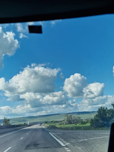 The image captures a landscape view from the perspective of a vehicle, likely through a side window, indicated by the dark frame on the far left. The scene is dominated by a bright blue sky filled with numerous large, fluffy white cumulus clouds that stretch across the upper and central parts of the frame. A small, blurry, dark rectangular object, possibly a sticker or reflection, is visible against the blue sky in the upper left section.

To the right, an asphalt road with clear white markings (a solid line on the edge and a dashed line for lanes) extends into the distance. Several vehicles, appearing as small white shapes, are visible on the road ahead. Along the roadside and in the mid-ground, green fields and rolling hills stretch towards the horizon, punctuated by trees and shrubs. Small white road markers are intermittently placed along the edge of the road.

The overall setting suggests a journey through a rural or semi-rural area, consistent with the provided location context of Barsukovskaya, Russia. The bright sunlight, clear blue sky, and distinct cloud formations indicate a sunny day, likely during late morning or afternoon. No specific people are visible, nor is any discernible text. The scene conveys a sense of open travel and natural beauty.