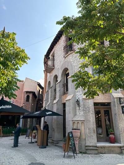 An outdoor shot shows a two-story building constructed from stone and brick, featuring arched windows with wrought-iron balconies. An open window box and decorative stone elements accentuate the façade. A doorway, framed by stone, leads into the building. Outdoor seating with umbrellas is set up on the cobblestone street. Two individuals wearing suits are observed, one standing and the other seated. Above, tree foliage partially obscures the building. The shot is taken in Domingo Arenas, Mexico.