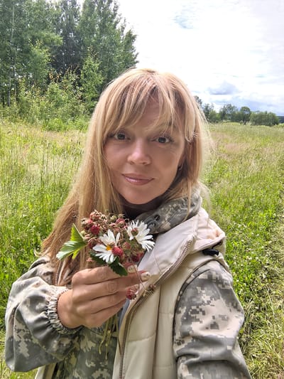 A medium shot captures a woman with blonde hair and bangs, holding a small bouquet of wild strawberries and white flowers in her left hand. She is wearing a camouflage jacket under a light-colored vest. The background consists of a lush green field with tall grass and wildflowers, bordered by trees. The sky is partly cloudy with patches of blue. The image appears to be taken outdoors in a rural or natural setting, possibly in Basharovo, Russia.