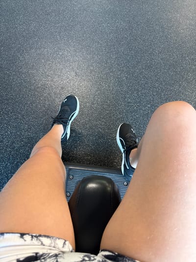 This is a high-angle, full-body shot of a person sitting down on a black padded bench. The person is wearing black Puma sneakers with white accents and black and white patterned shorts. Their legs are bare and lightly tanned. The bench appears to be part of a gym machine, as indicated by the textured black floor and the metal footplate visible under the person's feet. The lighting suggests an indoor environment, possibly a gym. The exact location in the City of Yonkers, United States of America cannot be determined from the image alone.