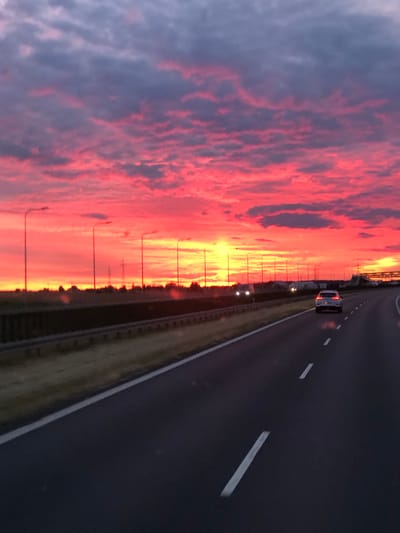 The image depicts a highway scene at sunset in Zaparcin, Poland. The sky is dominated by vibrant hues of pink and orange, with clouds casting dramatic shadows. A white SUV is driving on the right lane of the highway, while other vehicles are visible in the distance. Streetlights line the side of the road, and a guardrail separates the highway from the adjacent landscape. The overall impression is one of a scenic drive during a picturesque sunset.