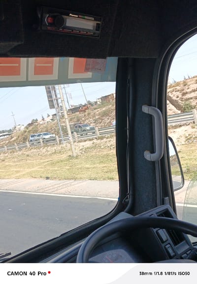 From the interior of a truck, a road stretches into the distance, flanked by grassy hillsides and modest buildings. Several vehicles, including a white SUV and a military-style truck, are visible on the road. A brown cow grazes on the hillside. The truck's dashboard and steering wheel dominate the foreground, and a Pioneer radio unit is mounted above the windshield. A gray grab handle is attached to the right pillar of the truck's cabin.