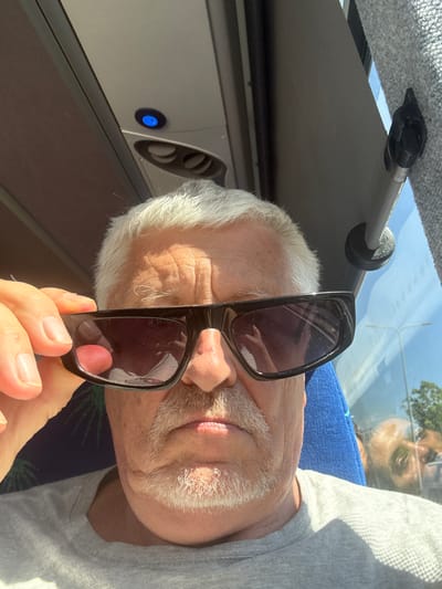A close-up, selfie-style image shows a middle-aged man with white hair and a grey mustache and goatee wearing sunglasses. He is holding the sunglasses down with his left hand, revealing part of his forehead and eyes. The man is wearing a grey t-shirt and appears to be sitting inside a vehicle, possibly a bus, as evidenced by the interior paneling and a window with a reflection. The lighting suggests it is daytime and sunny. The background shows the interior of a vehicle and a reflection of trees and sky in the window. The location is the city of Beer Tuvia Regional Council, Israel.