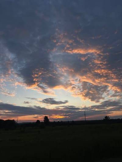 This is a vertical, outdoor, medium shot photograph of a sunset over a rural landscape. The sky is filled with dramatic clouds, some dark gray and others illuminated with vibrant oranges and pinks from the setting sun. Beams of sunlight pierce through the clouds, creating a dramatic effect. The horizon line is low, with a dark silhouette of trees and rolling hills. A single utility pole is visible on the right side of the frame, and a field of dark green grass occupies the foreground, completely silhouetted against the sky. The image appears to be taken from a distance, with no people or specific man-made structures visible beyond the utility pole and distant treeline. The overall mood is serene and atmospheric, capturing the beauty of twilight in a natural setting. The location is in the city of Sidorovy Gory, Russia.