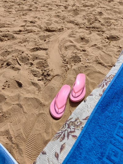 The image shows a pair of pink flip-flops lying on a sandy beach in Myadzyel, Belarus. The sand has numerous footprints imprinted on it, indicating recent activity. To the right of the flip-flops, a blue towel is placed on top of a patterned mat. The scene appears to be a relaxed setting, likely a sunny day at the beach, given the sand and the presence of beach accessories. There are no people visible in the frame.