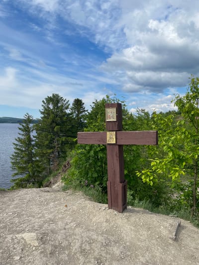 A prominent dark brown wooden cross stands outdoors on an elevated, rocky bank, overlooking a large body of water, likely a lake or river. Two religious icons are affixed to the cross's horizontal beam. The icon on the left depicts a figure with a halo, possibly a saint, rendered in a traditional style with a golden border. The icon on the right also features a haloed religious figure, possibly Christ Pantocrator or another saint, holding a scroll or book, set against a golden background.

Lush green foliage, including trees and bushes, densely frames the cross and the background. The expansive body of water stretches into the distance, meeting a hazy horizon or distant shore. Above, the sky is partly cloudy, displaying large grey clouds alongside patches of clear blue. The ground in the foreground consists of grey, rocky gravel, on which a small orange butterfly is visible in one of the close-up views.

The scene is captured during the daytime under natural light, suggesting a tranquil and possibly revered location. The setting, identified as Пятый километр, Russia, evokes a sense of peace and natural beauty, likely serving as a memorial or spiritual landmark. No discernable text is clearly visible on the icons themselves, but they clearly represent religious figures.