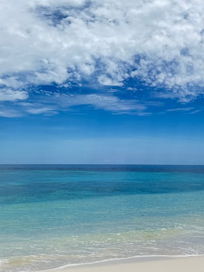 The image depicts a beach scene in Ukulhas, Maldives. The right side of the image shows a sandy beach with water washing ashore. The water transitions in color from a light, almost clear, color near the beach to darker shades of blue. The left side of the image displays a blue sky with scattered clouds.