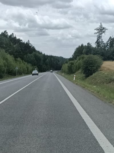 The image captures a section of a two-lane asphalt road in Čistá, Czechia. The road is marked with a white dashed line separating the lanes and a solid white line along the right shoulder. The scene is bordered by dense green trees and shrubs on the left and right, with a sparse grassy area and a hint of a dry field on the far right. The sky is overcast with heavy grey clouds.
In the distance, on the left lane, a dark-colored car is visible, appearing to be traveling away from the viewer. Further down the road, another lighter-colored car is also moving in the same direction. Roadside markers with orange and white tops are placed along the right side of the road. A triangular road sign is present on the left side of the road, near the edge of the treeline. The overall atmosphere suggests a cloudy day on a rural highway.