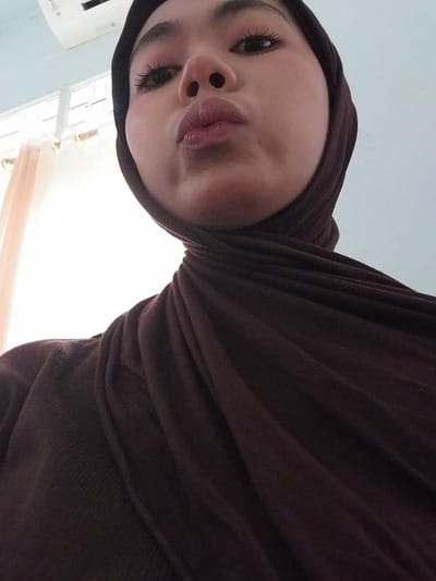 The media file is a close-up, low-angle selfie of a young woman wearing a dark brown hijab. The image is oriented with the woman's face appearing upside down. She is making a kissing face, with her lips pursed. Her eyes are open and looking towards the camera, and she is wearing mascara. The background is out of focus, but appears to be an indoor setting with a ceiling and light fixtures visible, suggesting it might be daytime. The woman's attire and the quality of light hint at a potentially warm climate. There are no discernible objects or text in the image. The setting seems to be a private indoor space, possibly a home. The overall mood of the image is playful and lighthearted, due to the woman's facial expression and the casual nature of the selfie. Given the provided location context of Watampone, Indonesia, the image likely depicts a moment in the daily life of a young woman in that region.