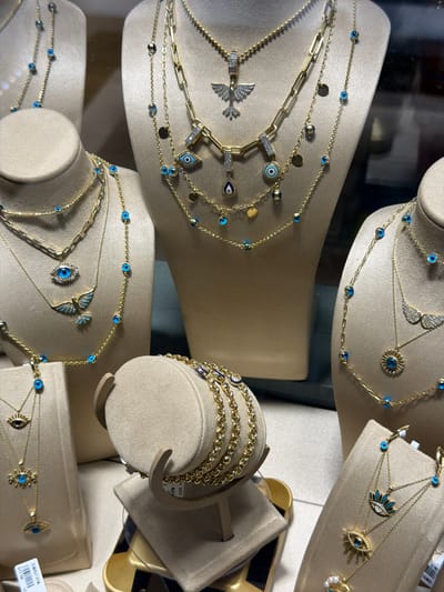 The image shows a display of gold jewelry featuring blue evil eye charms and other decorative elements. The jewelry is arranged on multiple mannequins and display stands, creating a layered presentation. The background appears to be a store window, possibly in Pendik, Turkey, as suggested by external context. Multiple necklaces, bracelets, and earrings are visible, all appearing to be made of gold and adorned with blue evil eye motifs, as well as some with bird and sun-shaped pendants. The lighting suggests an indoor setting, possibly a jewelry store.