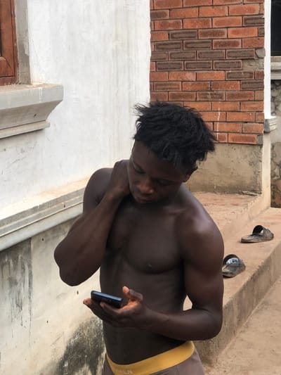 The scene, located in Oraukwu, Nigeria, depicts a shirtless young adult male with dark skin and dark, textured hair. He is oriented slightly left of the camera, looking down at a dark-colored smartphone held in his left hand. His right hand rests on the back of his neck. He wears brown or grey underwear with a yellow waistband. To the right of the subject, on a concrete step, are two dark, worn flip-flops. The background consists of a light-colored plastered wall with a partially visible wooden window frame on the left, and an exposed red and brown brick wall section on the right. The immediate ground surface is concrete.