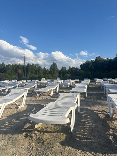 The image depicts a sandy area with numerous empty white plastic lounge chairs arranged in rows. The chairs are positioned on the sand, with some casting long shadows, indicating a sunny day. In the background, there is a dense line of trees. Further back, a volleyball net is visible, suggesting a recreational area. A lamppost stands to the left of the frame, and utility wires are strung across the sky. The sky is blue with scattered white clouds. The scene is located in Санаторный, Russia. There are no people visible in the image.