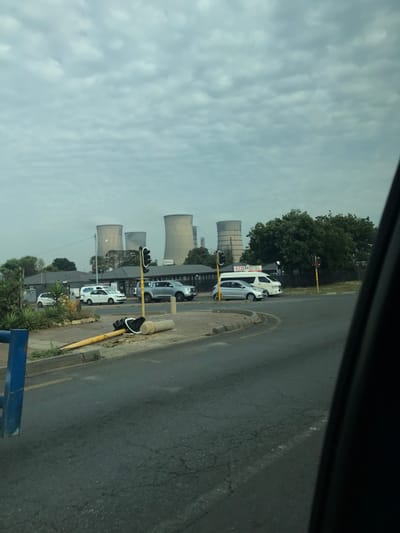 The image depicts an urban intersection in Kempton Park, South Africa, under an overcast sky. In the background, at least five large, light-colored cooling towers of a power station (likely Kelvin Power Station) are prominently visible above the treeline. The midground features a multi-lane road intersection with several vehicles, including white sedans, a silver pickup truck, and a white minibus taxi. Two upright traffic light poles, displaying red signals, are positioned on opposing corners of the intersection. Behind the vehicles and traffic lights, low-rise buildings and trees are present, with a sign clearly displaying "ECO MOTEL". In the immediate foreground, a yellow traffic light pole, detached from its base, lies horizontally on the paved curb area adjacent to the road, its signal head intact. The road surface exhibits cracking. No individuals are discernible, and vehicles appear to be in a state of transit or waiting.