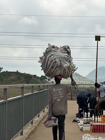 In Angwan Sayawa, Nigeria, a person walks with a large bundle of goods balanced on their head. The bundle is covered with a light-colored material, possibly plastic sheeting, tied with rope, and appears to be made up of compressed items. The person is wearing a long-sleeved, light-colored shirt with logos visible on the back, including "Cigna," "AT&T," "CITIZEN," "DAIAN," "ODDERBOX," and "FRESH." They are also wearing dark trousers and carrying a small pink and white patterned bag in their left hand.

The scene is outdoors, with a cloudy sky above. In the background, there are power lines and a utility pole with a rectangular fixture. To the left, a metal railing separates a pathway from a sloped, vegetated area with scattered trees and hills in the distance. To the right, other individuals are present, some appearing to be vendors with displays of various products, including bottles and containers, laid out on the ground. One person in a white shirt and another in dark clothing are visible among the vendors. The ground is paved, and a curb runs alongside the pathway.