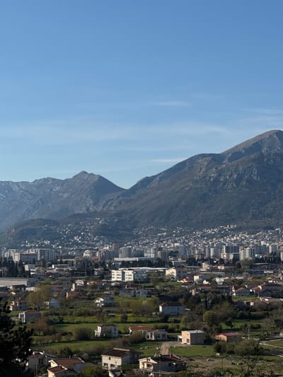 The image presents a mountainside urban landscape. A tall, rugged mountain range dominates the left side, with its rocky surface exposed, grading into greenery lower down. To the right, a densely built-up urban area stretches out, with a mix of high-rise apartment buildings closer to the mountain, and more dispersed residential houses further out. The sky is clear and blue on the left. The orientation of the scene has the sky on the left, the mountain in the center, and the urban area on the right. It is not possible to determine the location is Tomba, Montenegro with certainty.