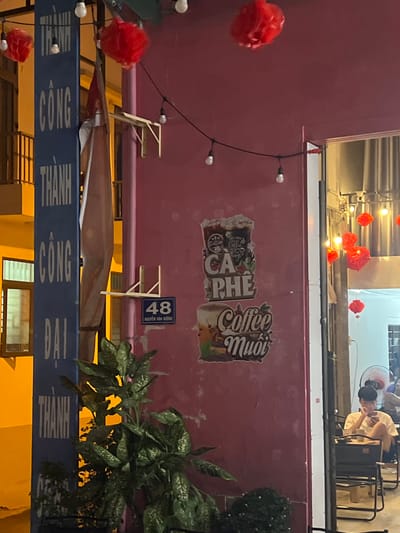 A street-level photograph captures a nighttime scene in Nam Nha Trang, Vietnam.
The primary focus is on a faded pink wall adorned with stickers reading "Cafe Phe" and "Coffee Muoi". A small blue sign with the number "48 Nguyen Van Huong" indicates the address. A string of small, bare bulbs hangs against the wall.
Below the wall is a blue sign displaying "THANH CONG BAI THANH". Red pom-pom decorations and tropical foliage add to the scene.
Above, partially visible through an opening, are glimpses of people seated in a cafe. The overall lighting suggests a warm, urban environment.