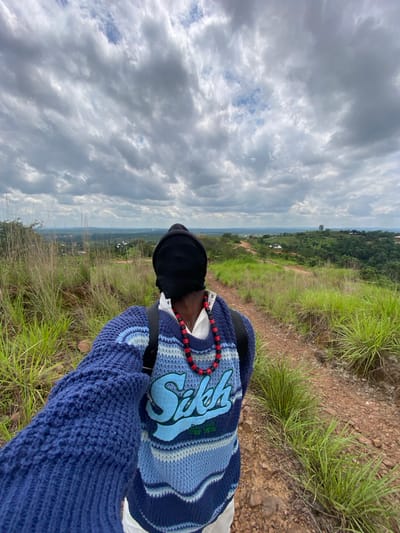 A person wearing a blue and white striped knit sweater with a blue logo on the back and a black head covering is standing on a dirt path in a grassy, hilly landscape. The person is holding up their right arm, as if taking a selfie. In the background, there is a vista of green hills, scattered buildings, and a cloudy sky. The path is reddish-brown and winding, with dry grass and some rocks on either side. The overall scene suggests an outdoor, natural environment, possibly on the outskirts of Ufueseni, Nigeria.